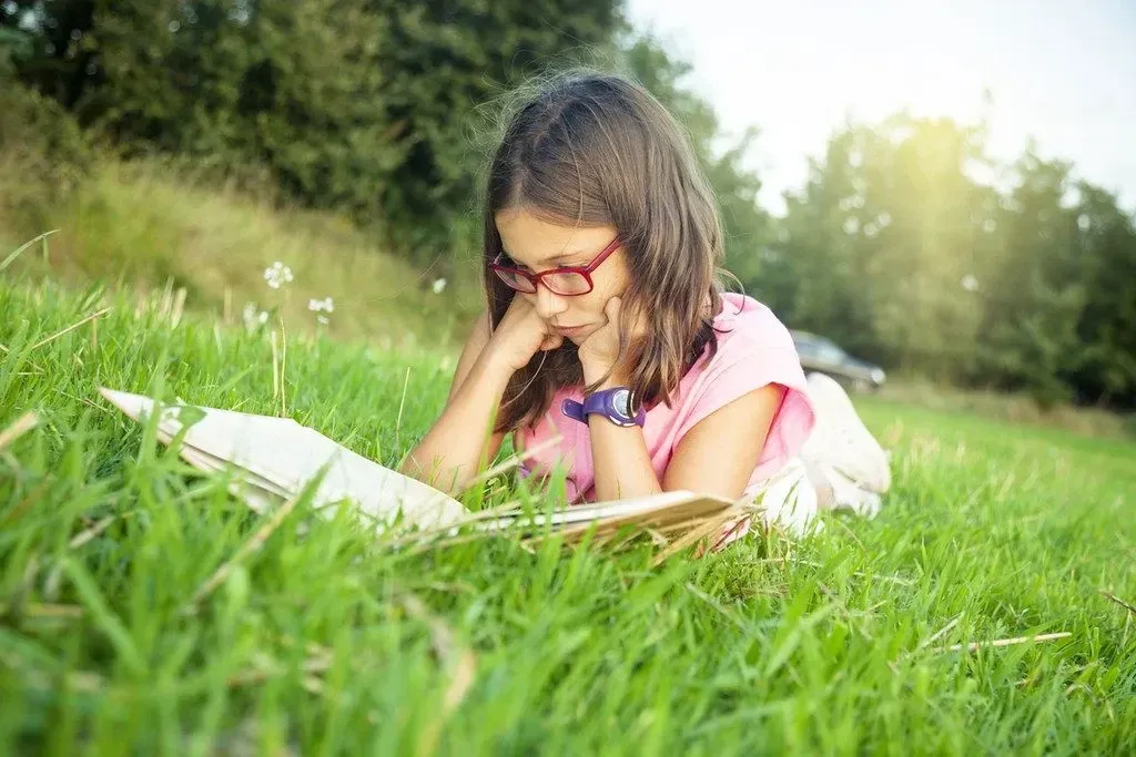 A tween girl reading a book on the grass.