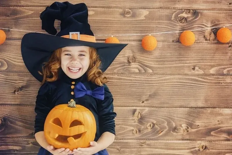Little girl dressed as a witch standing against a wooden background, smiling and holding a Jack O' Lantern. 