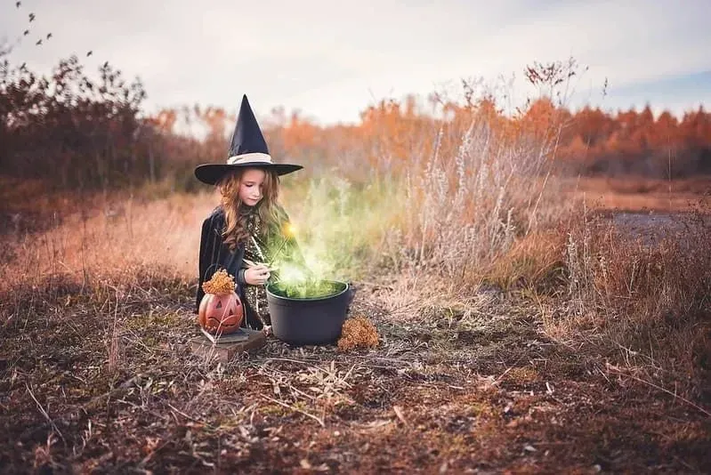 Little girl dressed as a witch sitting in the middle of a field with a cauldron that's emitting green steam.