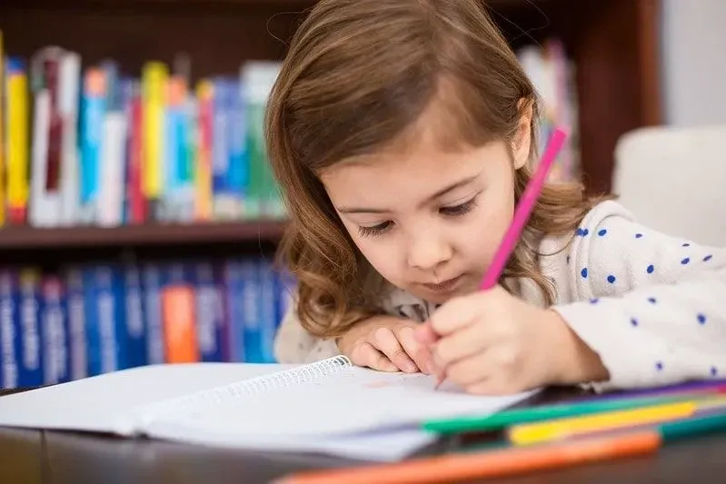 Little girl sitting at a desk writing in her exercise book with colouring pencils.