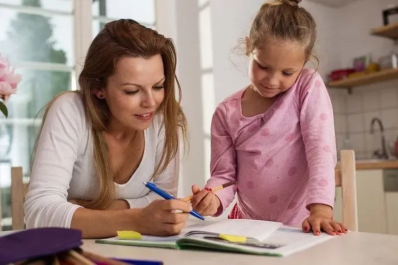 Mum sat at table helping her daughter with her homework.