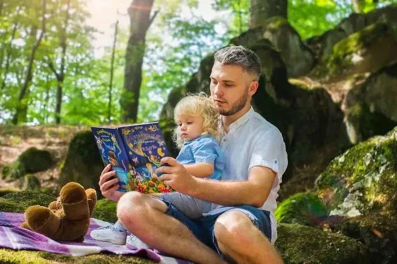 A man is sat outdoors in a shaded woodland area, reading to his son on his lap.