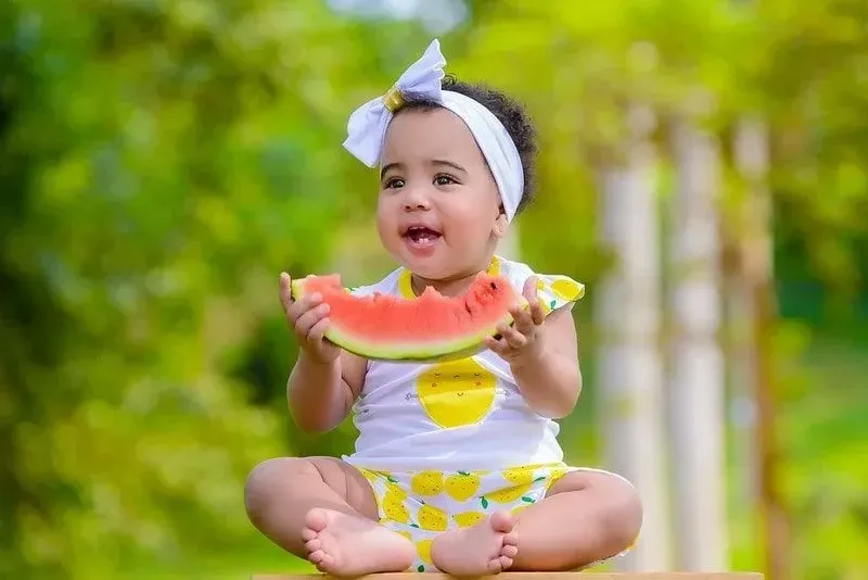 Toddler wearing a bow headband eating a piece of watermelon.