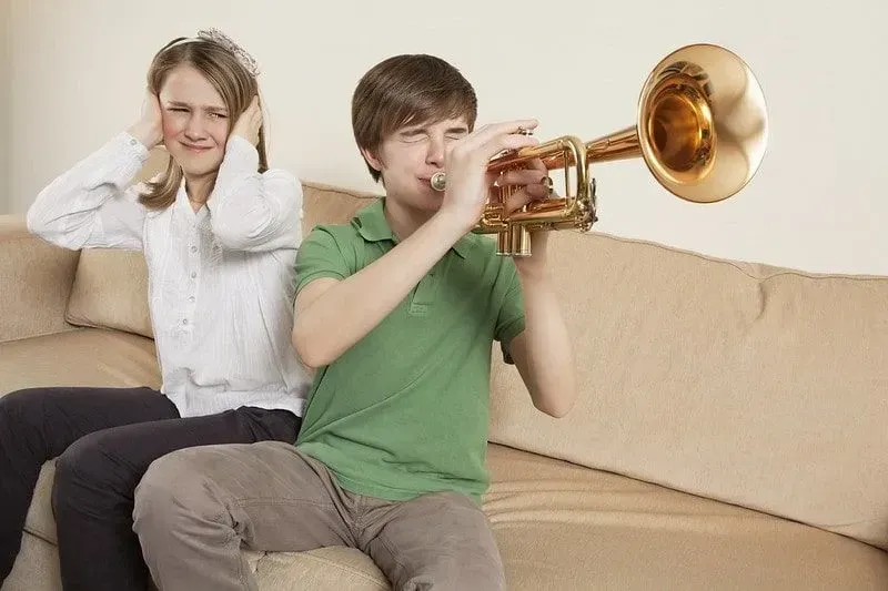 Boy plays the trumpet and his sister sat next to him covers her ears. 