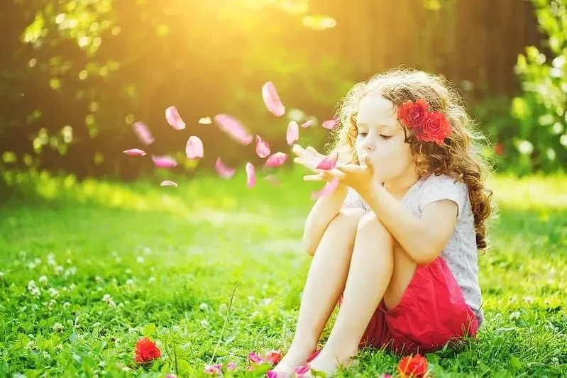 Little girl sat on the grass blowing flower petals out her hands.