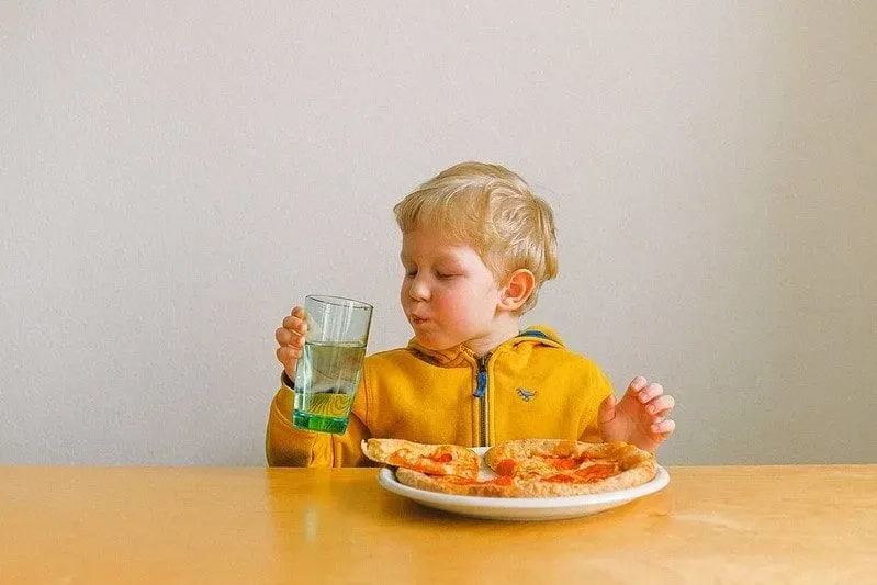Young boy sat at the table eating pizza and drinking water.