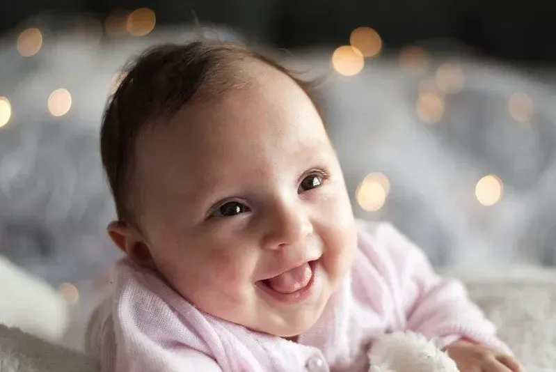 Happy baby girl, wearing a pink baby grow, with lights shining behind her.