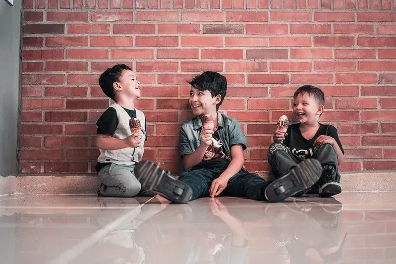 Three boys sitting on the floor by a wall eating ice cream and laughing together.