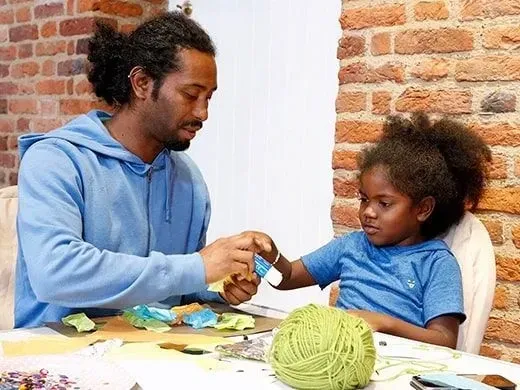 Father and daughter sat at the table doing crafts together. Dad in handing his daughter glue.