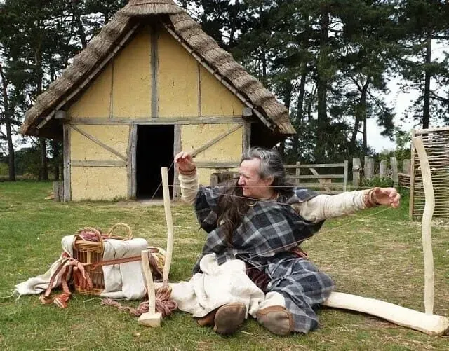 Woman dressed in Anglo-Saxon clothing, in an Anglo-Saxon village, sat on the floor weaving.