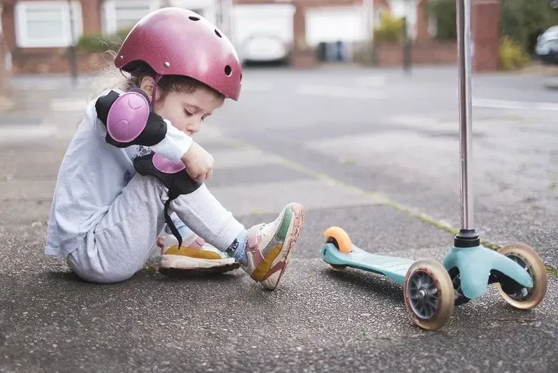 A little girl is putting on kneepads before going on her scooter.