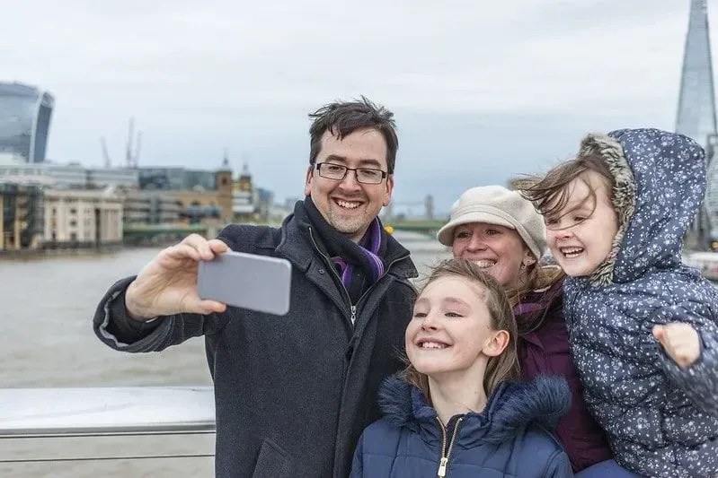 Family taking a selfie by the Thames river with London monuments in the background.