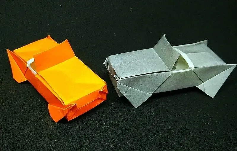 Two origami cars, one orange and one grey, on a black surface.