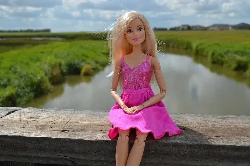 Barbie doll in a pink shiny dress sitting on the railing of a bridge, posing.