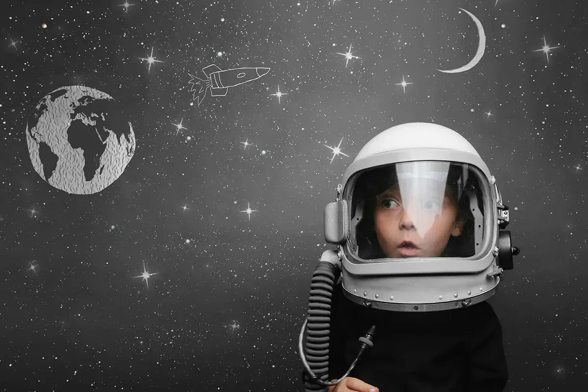 Young boy wearing an astronaut helmet standing in front of a blackboard with space drawings on it.n