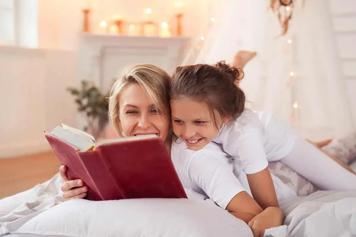A mother and daughter are lying down reading a book together smiling, learning about idioms.