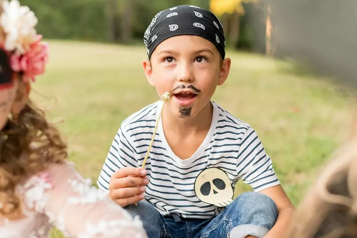 Boy dressed as a pirate, with a moustache and goatee drawn on and a bandana on his head, sat on the floor outside.