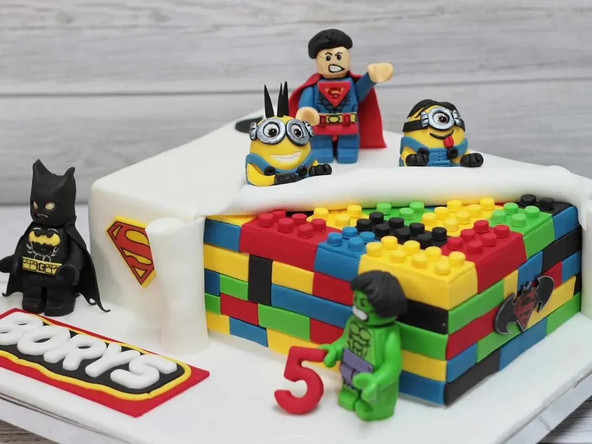 A colourful rectangular LEGO cake, part covered in fondant icing, with fondant LEGO superheroes including superman and batman placed on top of or around the cake.