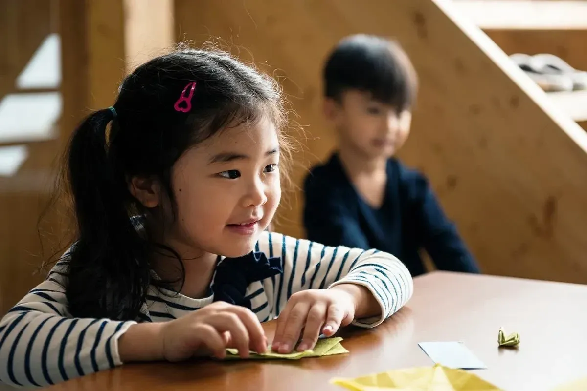 Little girl sat at the table smiling as she folds paper to make an origami squirrel.