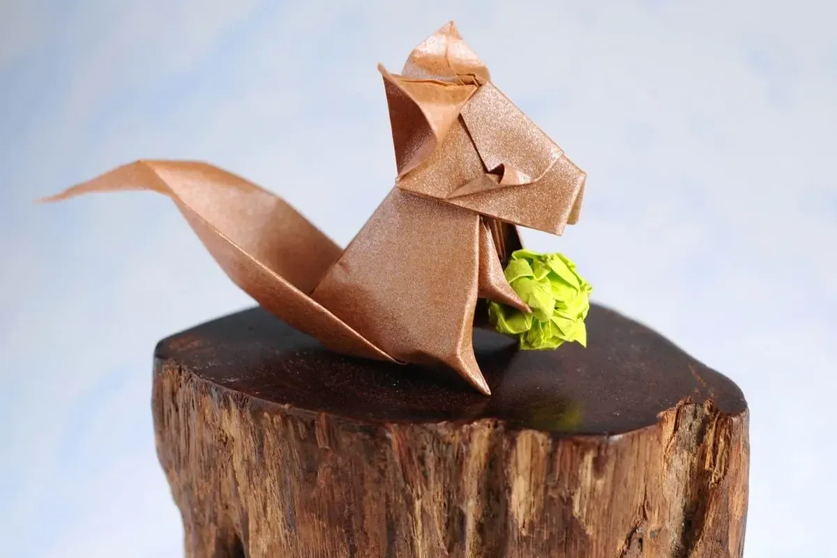 Shimmery brown origami squirrel placed on a polished log holding a green origami acorn.