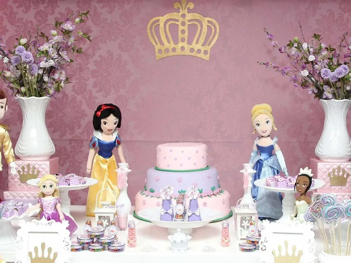 Pink and purple layered princess cake on a display table, surrounded by flowers, Disney princess toys and sweets.