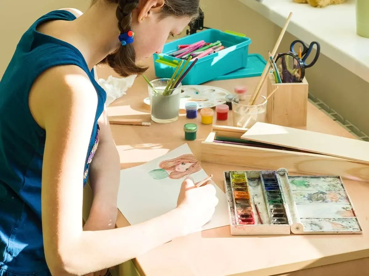 A teenage girl sits surrounded by art equipment, she is concentrating on completing a watercolour painting.