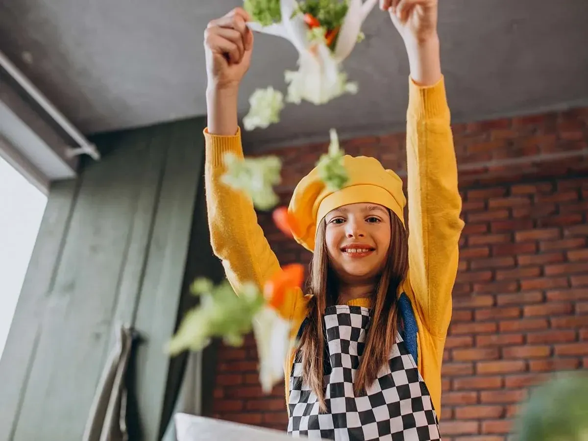 A tween girl wearing a chef's hat and apron tosses salad into the air whilst smiling at the camera.