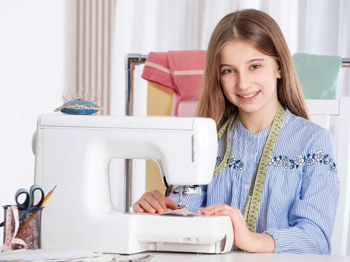 A tween girl sits at a desk in front of a sewing machine, she is surrounded by sewing equipment including a pin cushion and tape measure and is smiling at the camera.