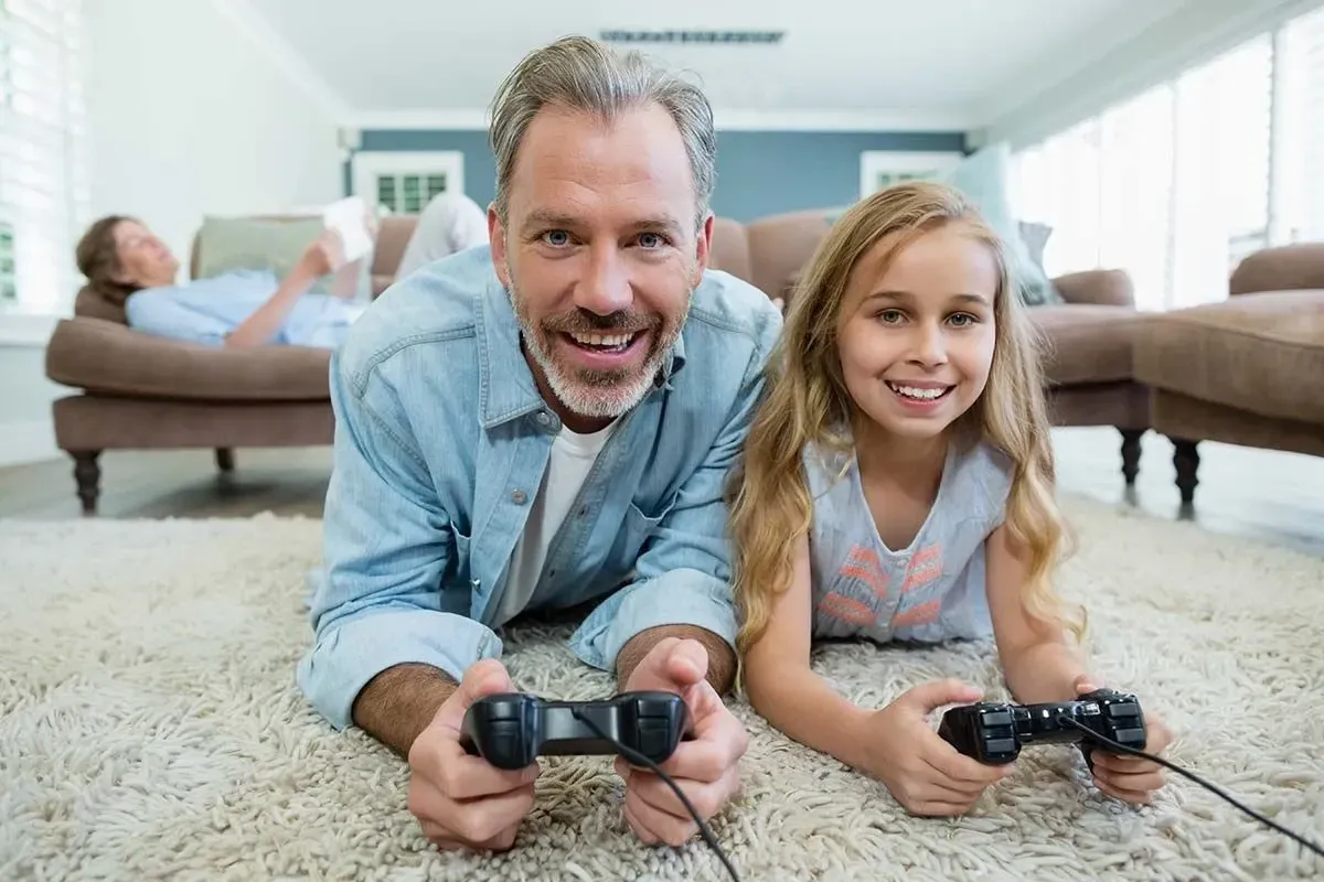 A girl and her father smile into the camera, they are enjoying her reward of playing a video game. 