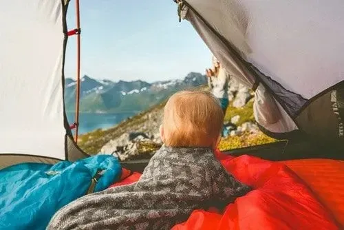 Toddler, wrapped in a blanket, lying on its tummy in the camping tent looking out the door at the view of mountains.