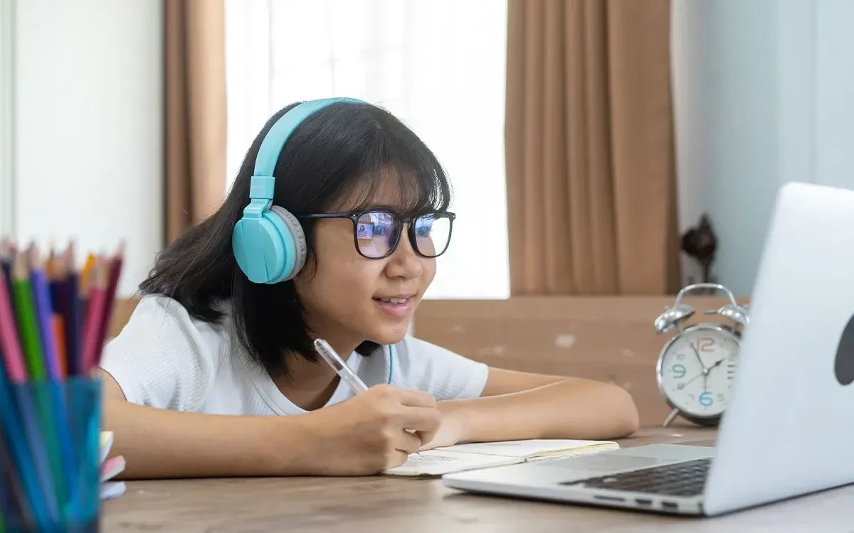 A girl wearing headphones and glasses using a laptop and notebook to learn about Roman numerals.