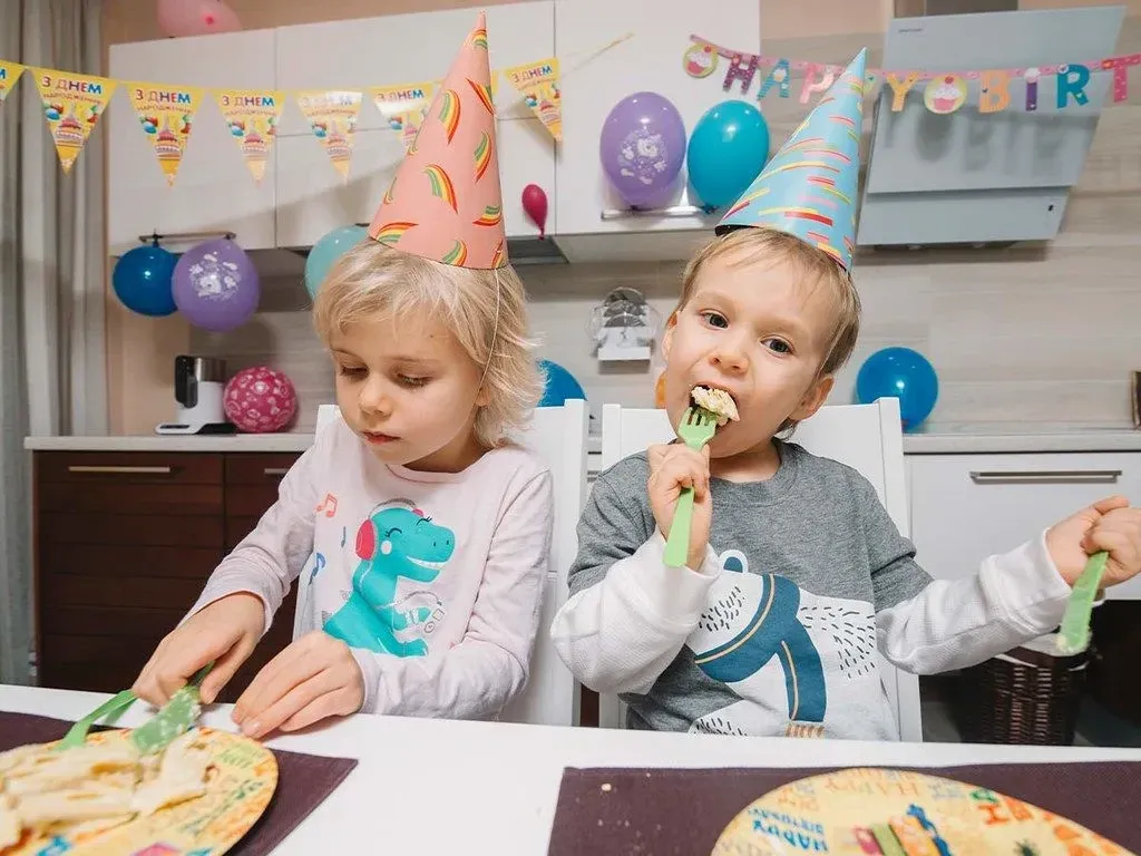 Two kids wearing party hats, enjoying a slice of cake at a birthday party.