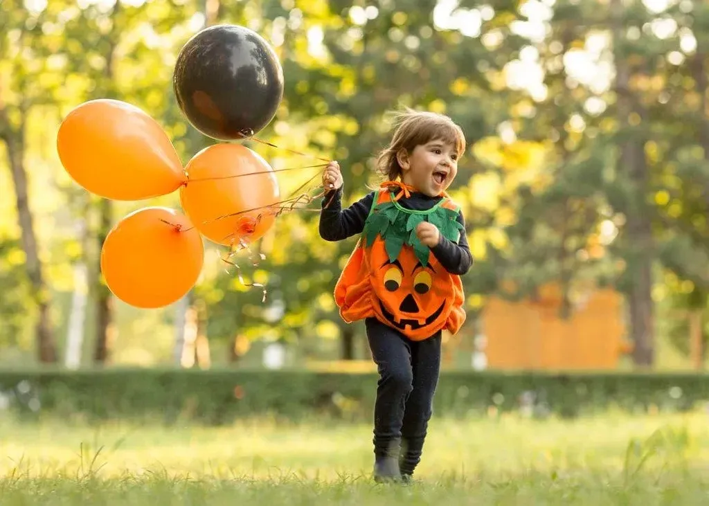 A child dressed in a Halloween costume holding orange and black balloons.