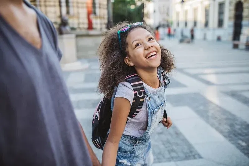 A young girl wearing denim kids' dungarees laughs as she walks with a parent.