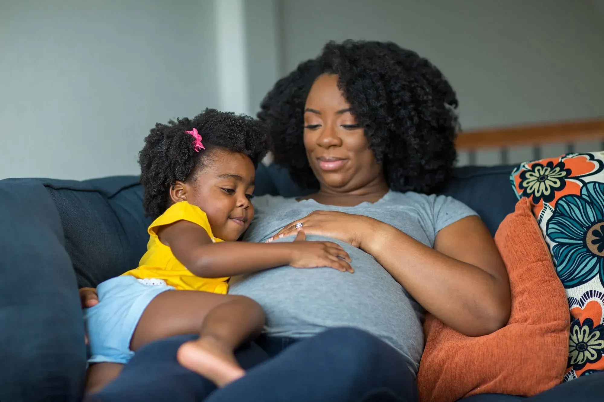 A pregnant mum sitting on the sofa with her toddler, both feeling her tummy. Image