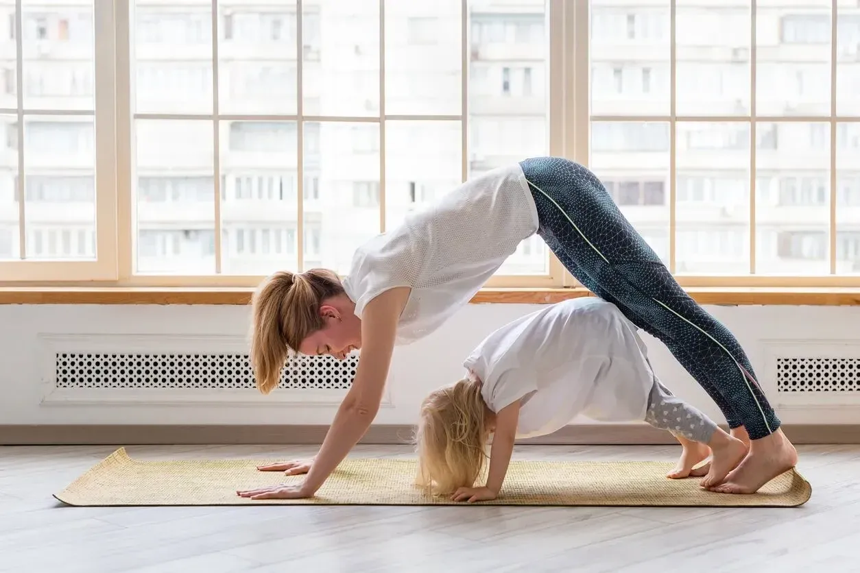 A mum and young daughter doing yoga together in a bright lit room with lots of windows.