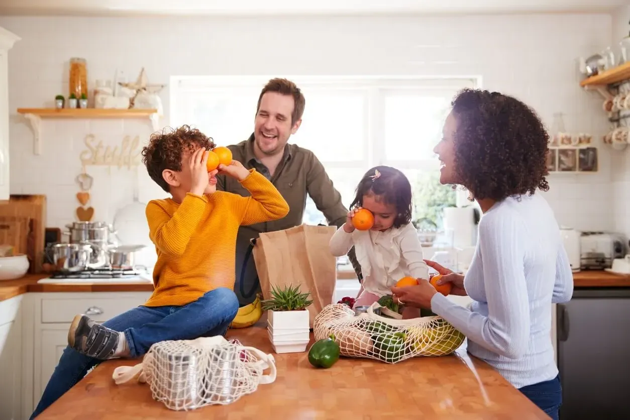 A family of parents and two kids laughing and unpacking groceries in the kitchen together. Image