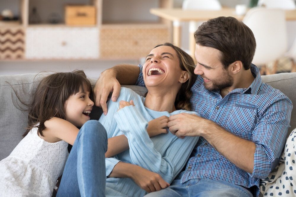 Cheerful people sitting on couch in living room have fun little daughter tickling mother laughing together with parents enjoy free time playing at home.