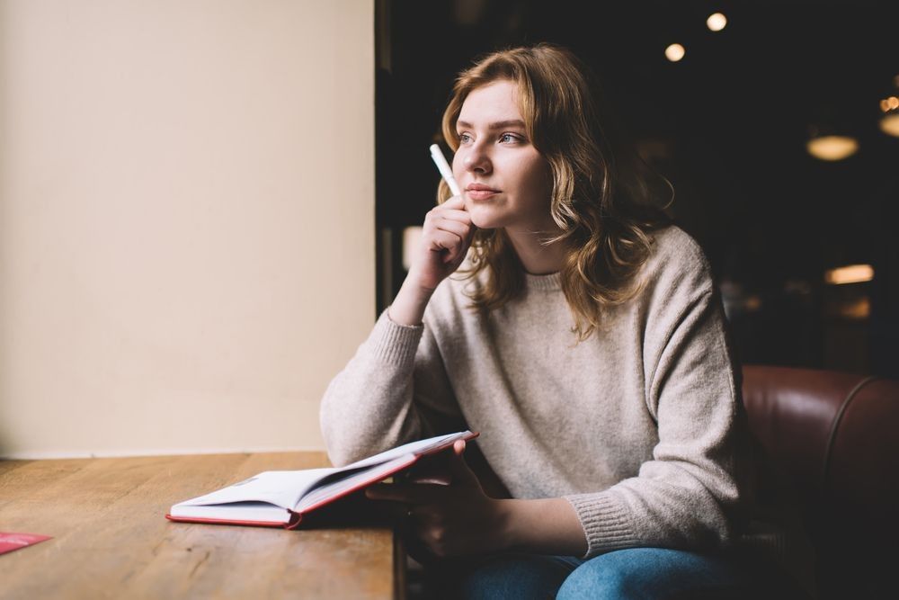 Young female novelist in baggy sweater thinking hand at chin and looking away through window while sitting at cafe table with open diary and pen in hand