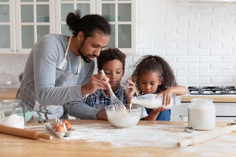 Happy caring young father teaching small adorable kids son & daughter mixing flour with milk.