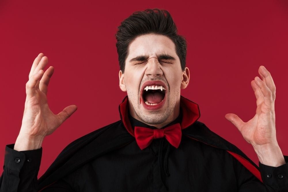 Image of angry vampire man with blood and fangs in black halloween costume screaming isolated over red wall.