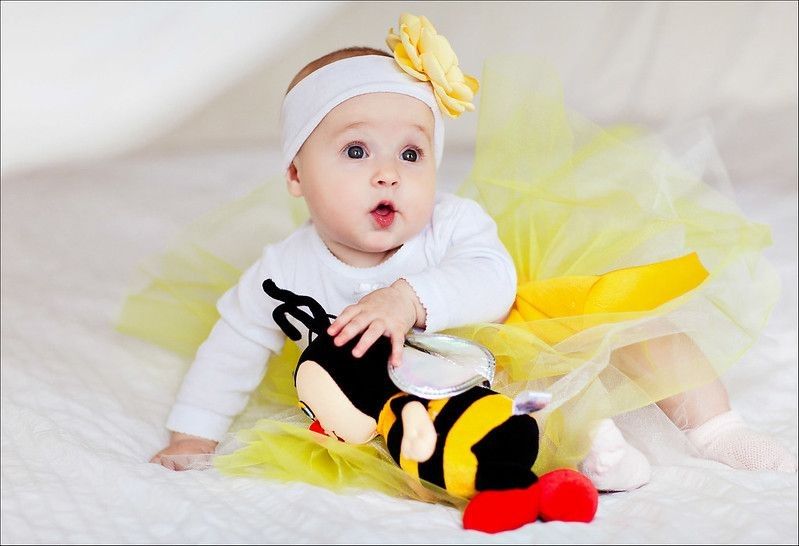 baby girl wearing white and yellow flower headband sitting on bed with bee toy