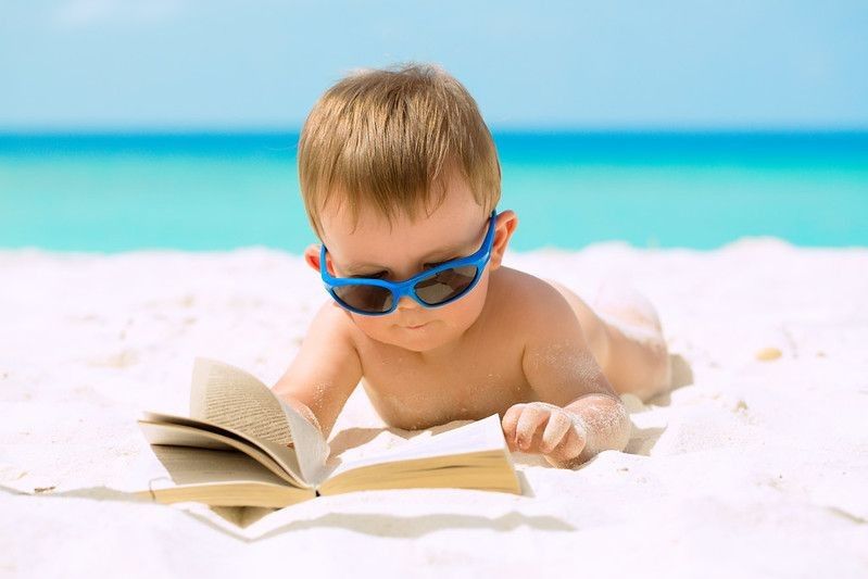 Cute baby boy with sunglasses lying on white sandy beach reading book