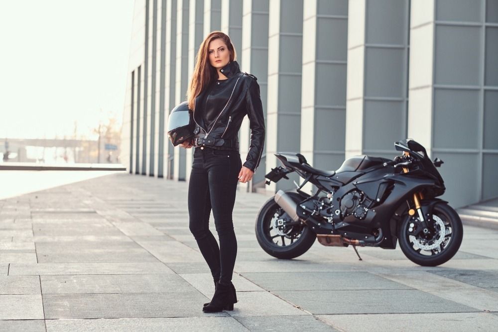 A beautiful biker girl holding helmet next superbike outside a building.