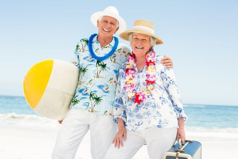 Senior couple wearing Hawaiian shirts holding surfboard on a beach.