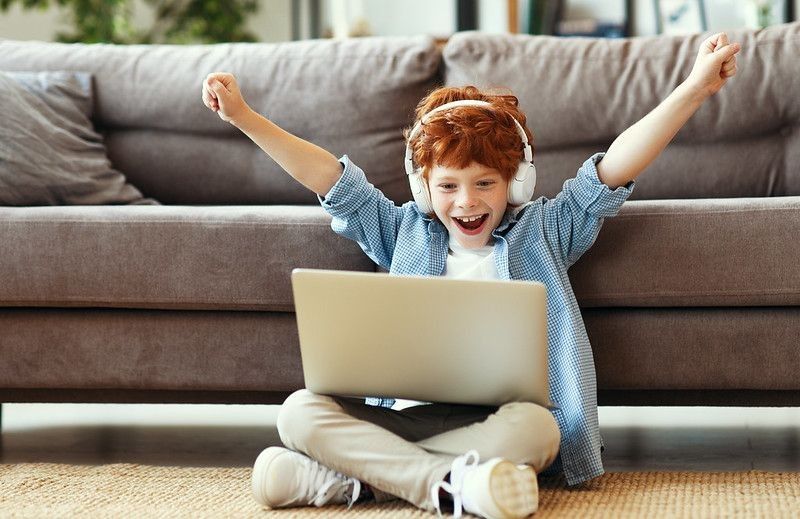Young boy in headphones raising arms and smiling while sitting crossed legged on floor and celebrating victory in video game on laptop