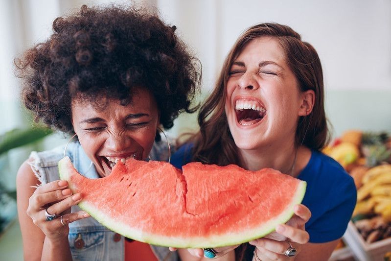 Female friends eating a watermelon slice and laughing together.