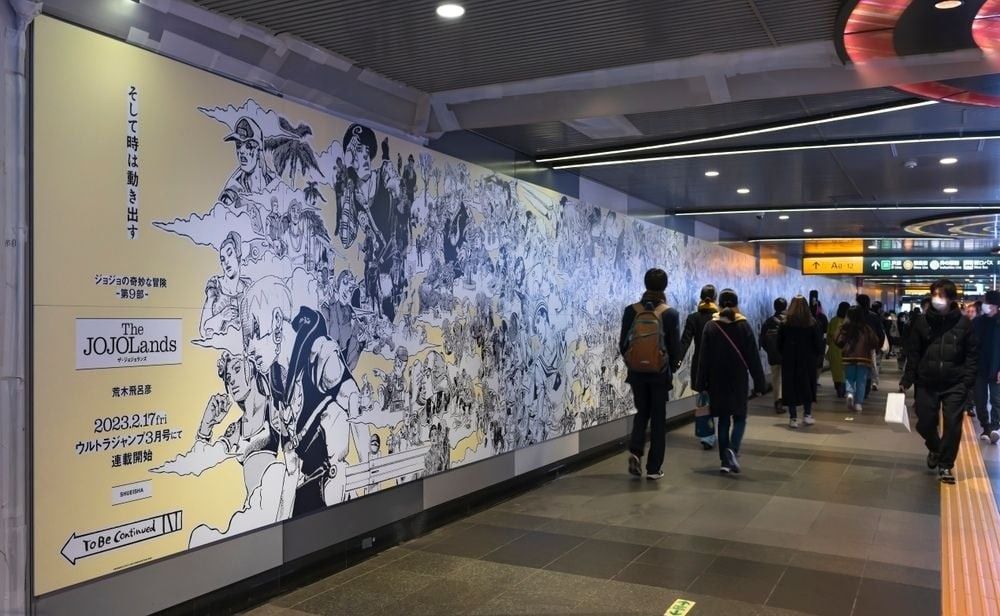 Travellers walking in the corridor of Shibuya Station along a giant poster depicting all the characters of the Japanese manga by Hirohiko Araki, JoJo's Bizarre Adventure.