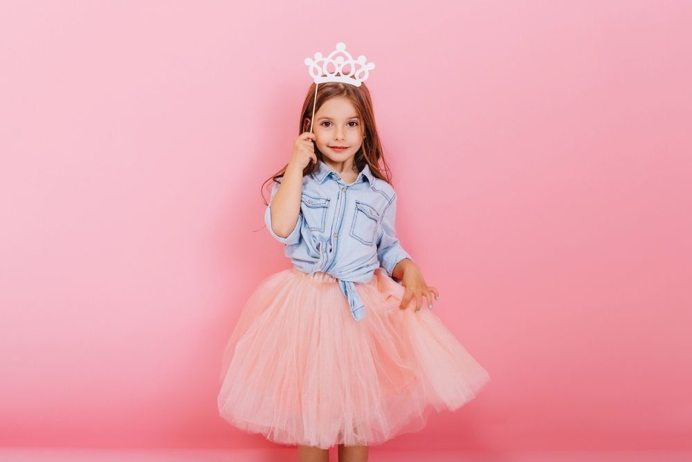 Cheerful little girl with long brunette hair in tulle skirt holding princess crown on head isolated on pink background. 