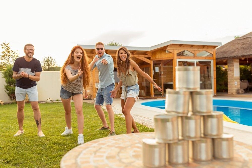 Group of cheerful young friends having fun playing the knock down tin cans by throwing a ball game while at poolside summertime outdoor party.
